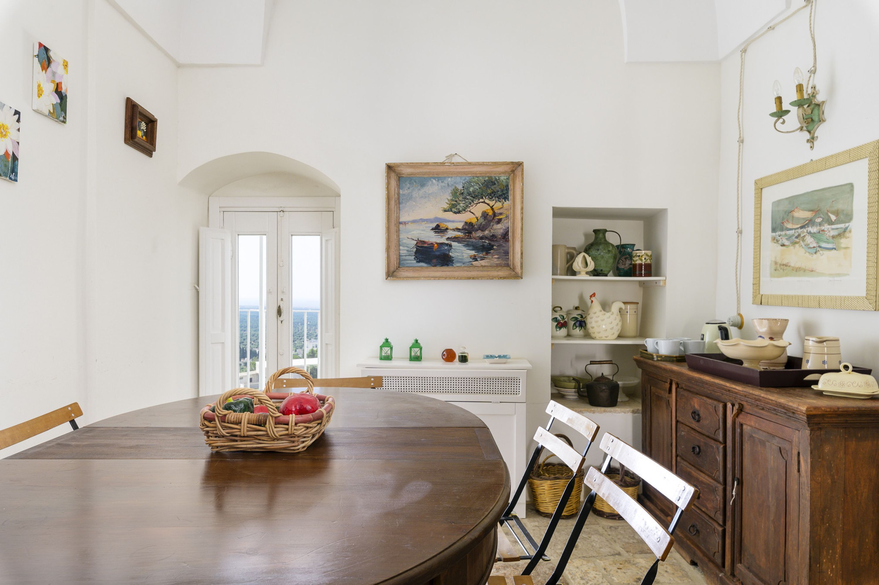 A dining area with a wooden table, chairs, a basket of fruit, and a painting on the wall.