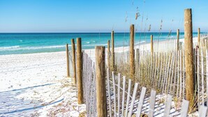 Une plage bordée d’une clôture en bois et d’herbes de plage, qui mène à une étendue de sable avec l’océan en toile de fond.