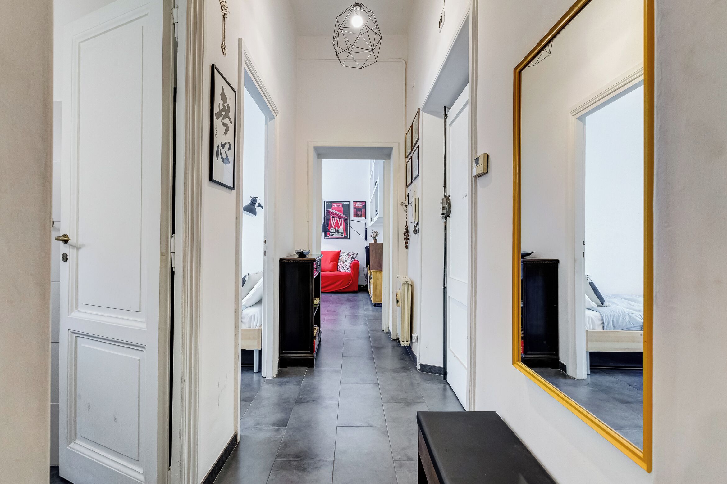 A hallway with a mirrored wall, a red sofa, and a bed in the background.
