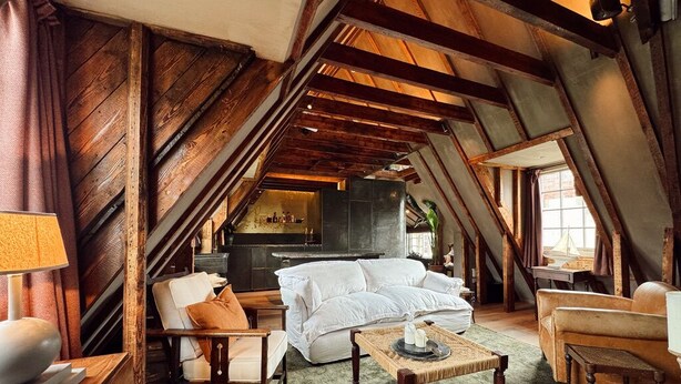 A cozy attic room with exposed wooden beams, a white sofa, a wooden chair, and a small table with candles.