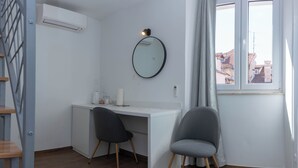 Ένα μοντέρνο δωμάτιο με ένα λευκό γραφείο, δύο καρέκλες, έναν στρογγυλό καθρέφτη και ένα παράθυρο με θέα σε κτίρια.