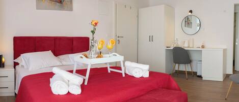 A hotel room with a red bedspread, white towels, a small table with orange drinks, a vase with flowers, and a desk with a chair.