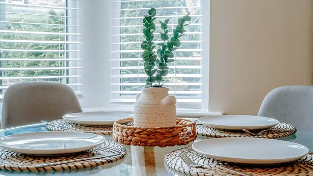 A dining table with plates and a vase of green plants, set against a window with blinds. - Stay Harrogate 3 Bed Holiday Home | Walk to Convention Centre | Parking (North Yorkshire)