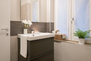 Une salle de bain moderne avec un lavabo blanc, un meuble sombre et un miroir au-dessus. On aperçoit une petite plante sur le rebord de la fenêtre et un panier rempli de serviettes.
