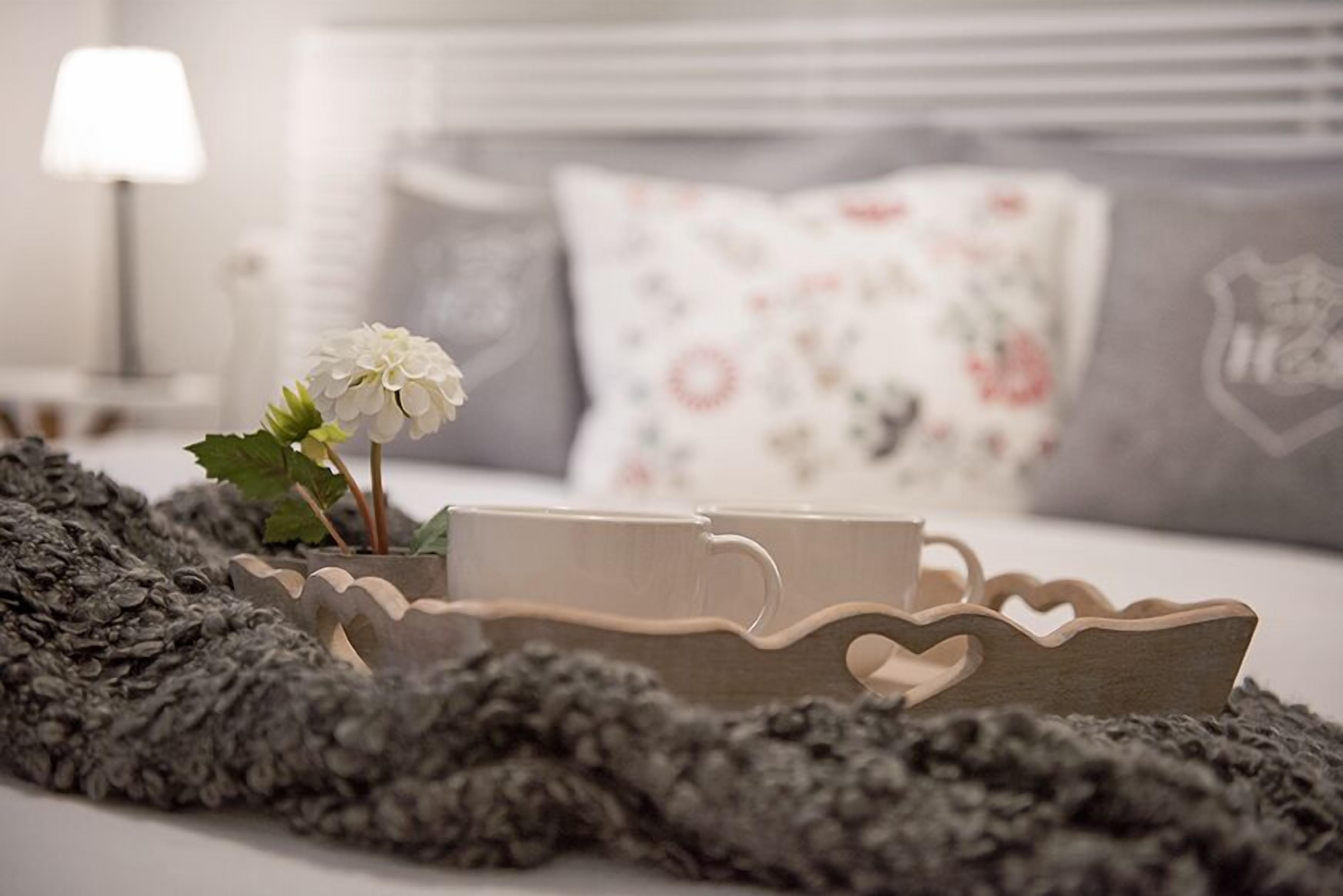 A cozy bedroom with a bed, a bedside lamp, a decorative pillow, and a tray with a cup and a flower.