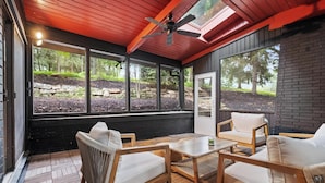 A covered patio with wooden furniture, large windows, and a ceiling fan.