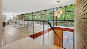 A spacious living area with a high ceiling, large windows, and a staircase with a wooden finish.