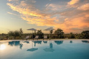 Ein Schwimmbecken mit Liegestühlen und Sonnenschirmen, umgeben von Bäumen und wolkenlosem Himmel.