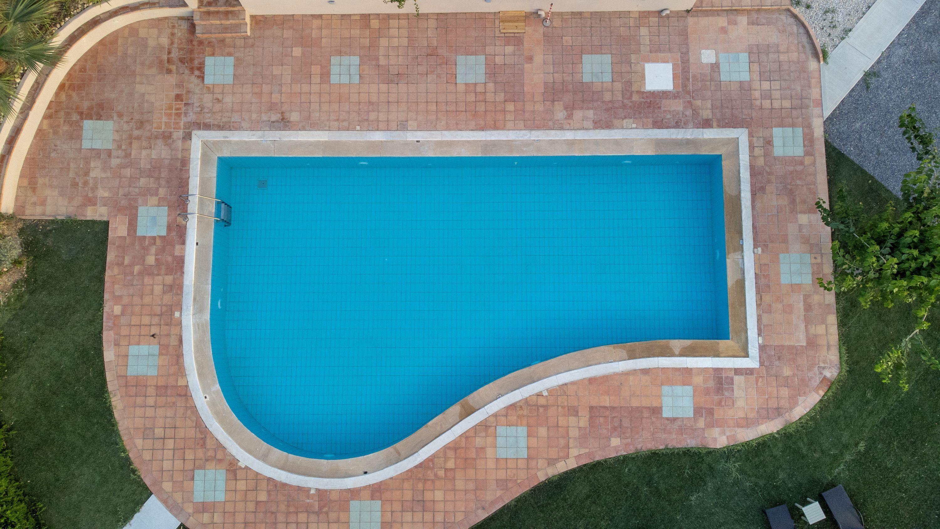 Vista aerea di una piscina rettangolare con bordo curvo, circondata da un solarium piastrellato con un mix di piastrelle marroni e beige.