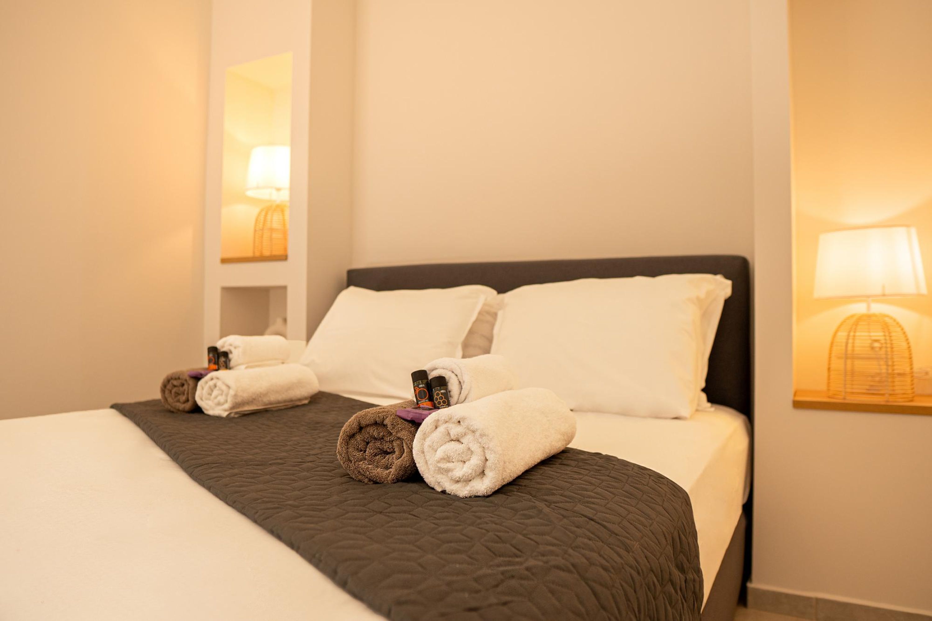 A neatly made bed with a dark quilt, two white pillows, and rolled towels on the side.
