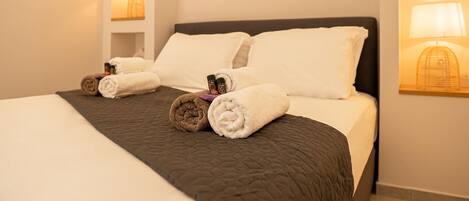 A neatly made bed with a dark quilt, two white pillows, and rolled towels on the side.