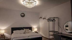 A bedroom with a bed, bedside lamps, a desk, a chair, a mirror, a coat rack, and a chandelier.