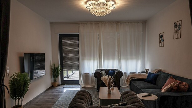 A modern living room with a dark sofa, a coffee table, and a TV on the wall. There is a large window with curtains, a potted plant, and a chandelier.