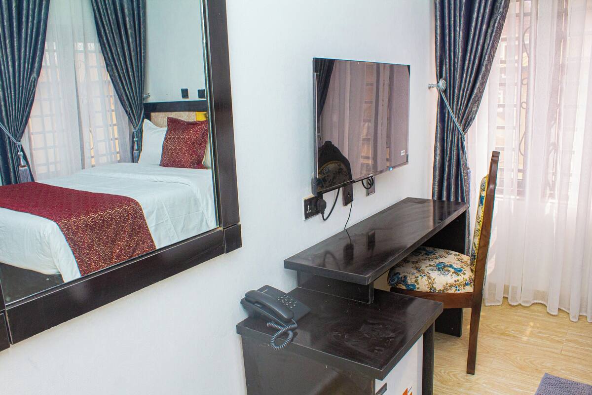 Habitación de hotel con cama, escritorio con teléfono, sillón tapizado con motivos florales, televisor de pantalla plana montado en la pared y un espejo grande que refleja la habitación.