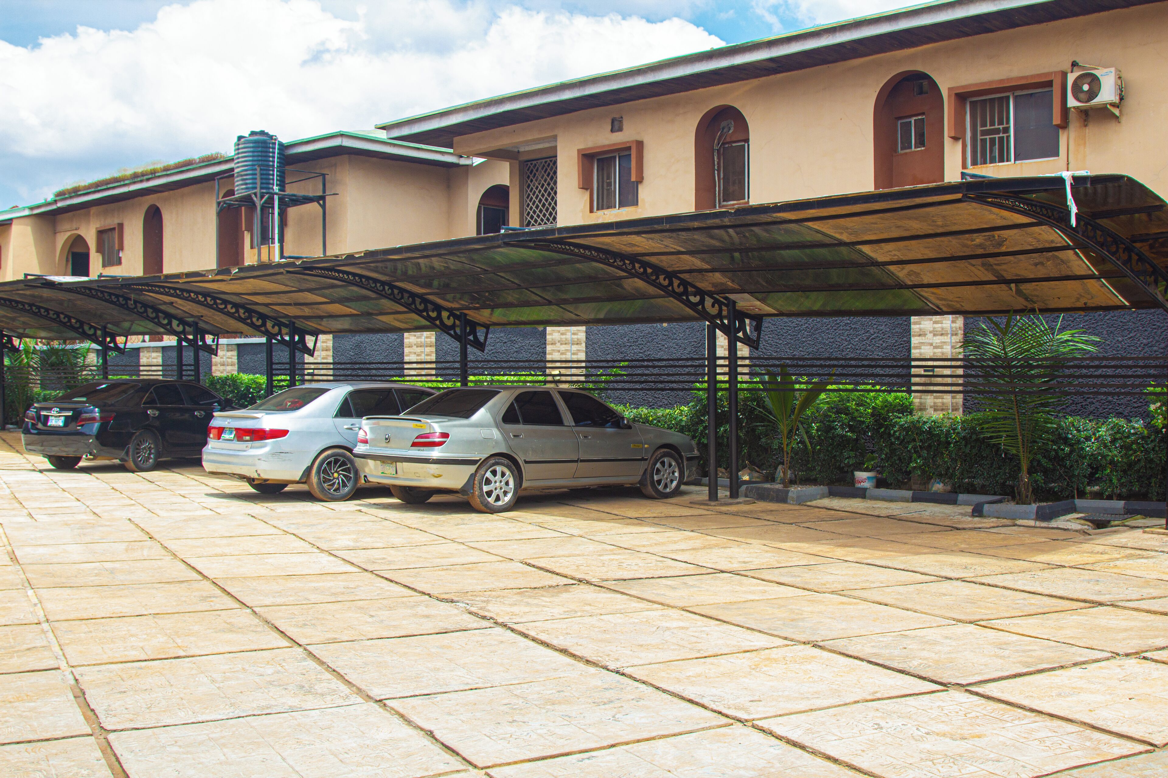 A parking area with two cars, a covered walkway, and a building with a water tank and arched windows.