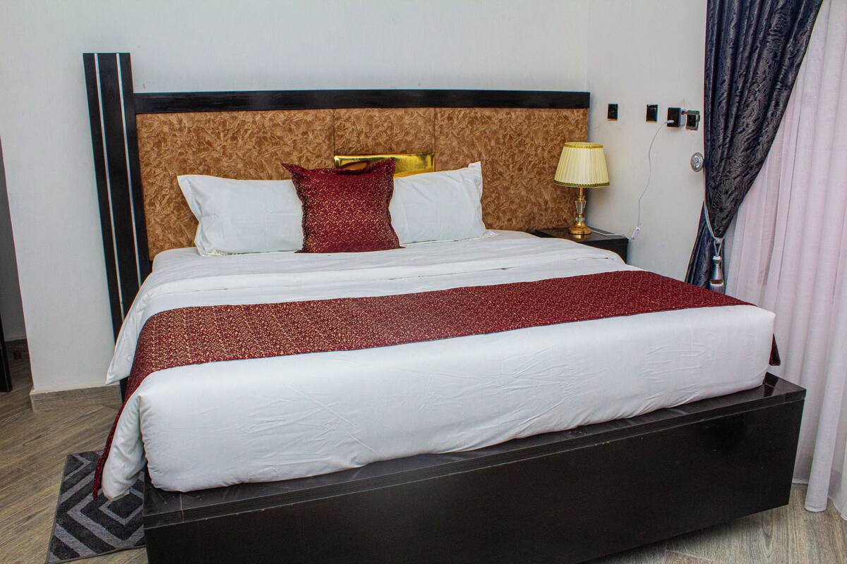 Una habitación de hotel con una cama grande, cabecera con diseño de madera, mesita de noche con lámpara y cortina en la ventana.