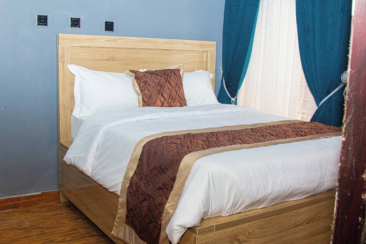 Una cama bien hecha con cabecera de madera, ropa de cama blanca y marrón, y una colcha estampada.