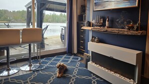 Una habitación con un ventanal grande que da a una masa de agua, una zona de estar con dos sillones, un perro y una estantería con adornos.