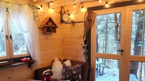 A cozy cabin interior with a wooden interior, a sofa with cushions, a bench, and a window overlooking a snowy forest.