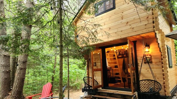 A wooden cabin with a porch, red Adirondack chairs, and a hanging basket.