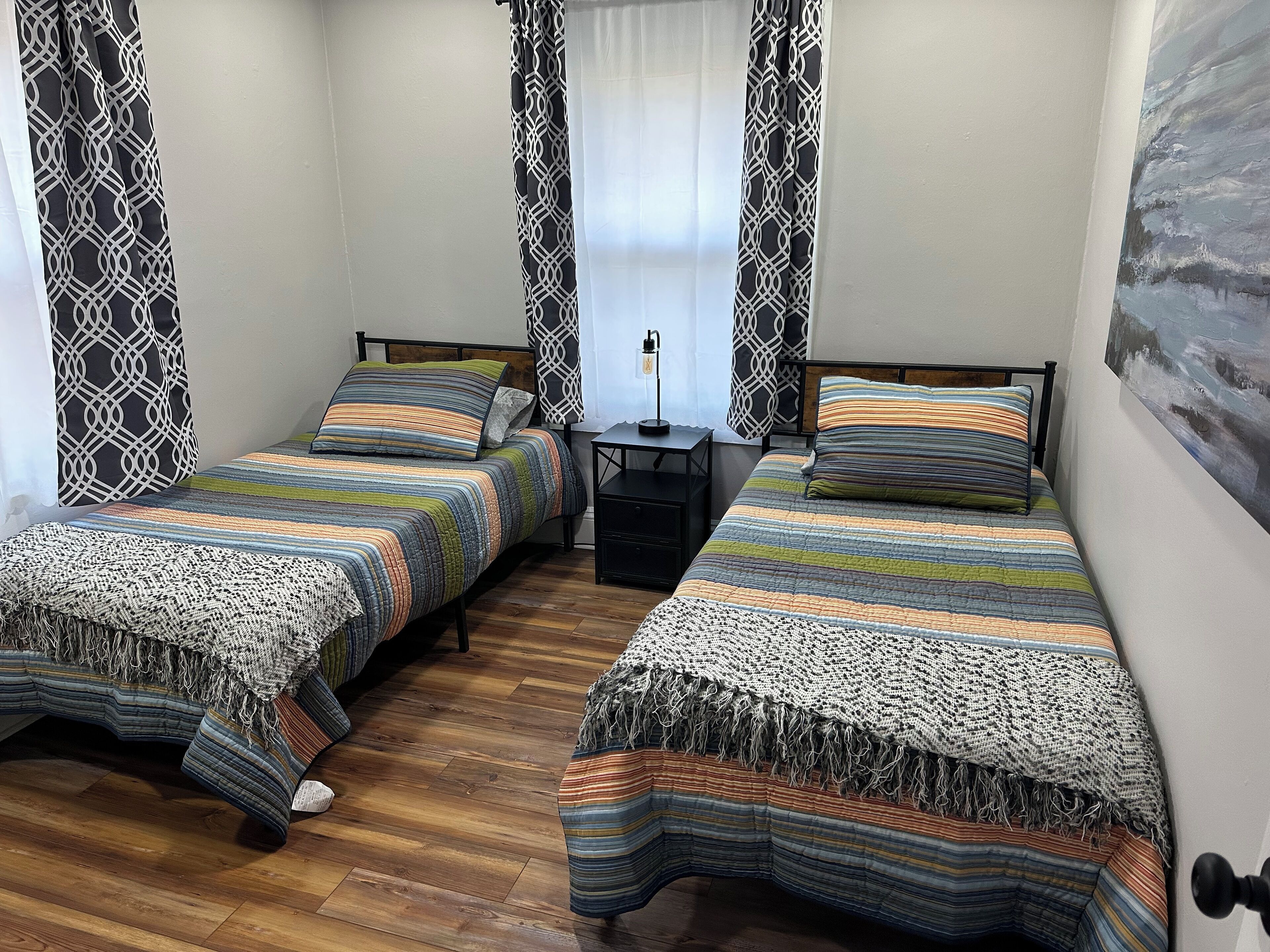 A room with two beds, each with a striped bedspread and a woven throw blanket. There is a small table with a lamp between the beds, and a painting on the wall.