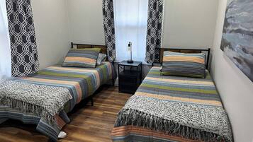 A room with two beds, each with a striped bedspread and a woven throw blanket. There is a small table with a lamp between the beds, and a painting on the wall.