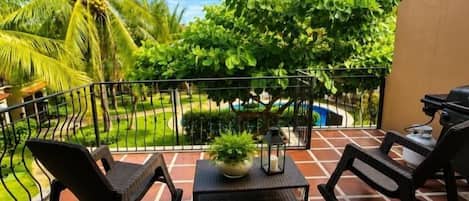 Un balcón con dos mecedoras, una mesita con una planta, y vista a una piscina y follaje.