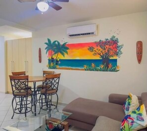A living room with a dining table, chairs, a sofa, and a colorful mural of a sunset and palm trees.