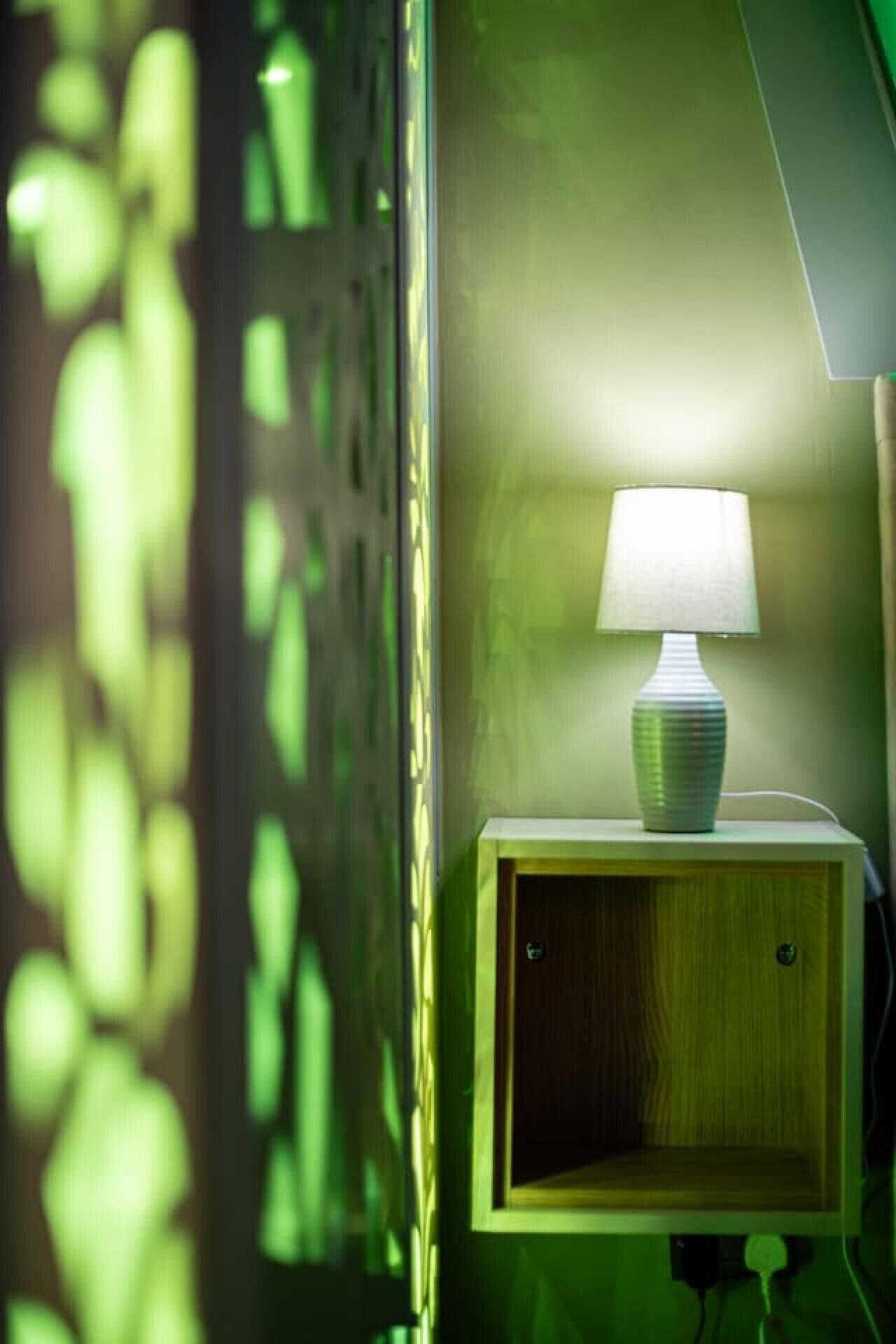 A bedside table with a lamp, a wooden cabinet below, and a green wall.