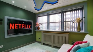 Une pièce avec une grande télévision affichant le logo « NETFLIX », un radiateur, une fenêtre avec des stores et un canapé blanc agrémenté de coussins colorés.