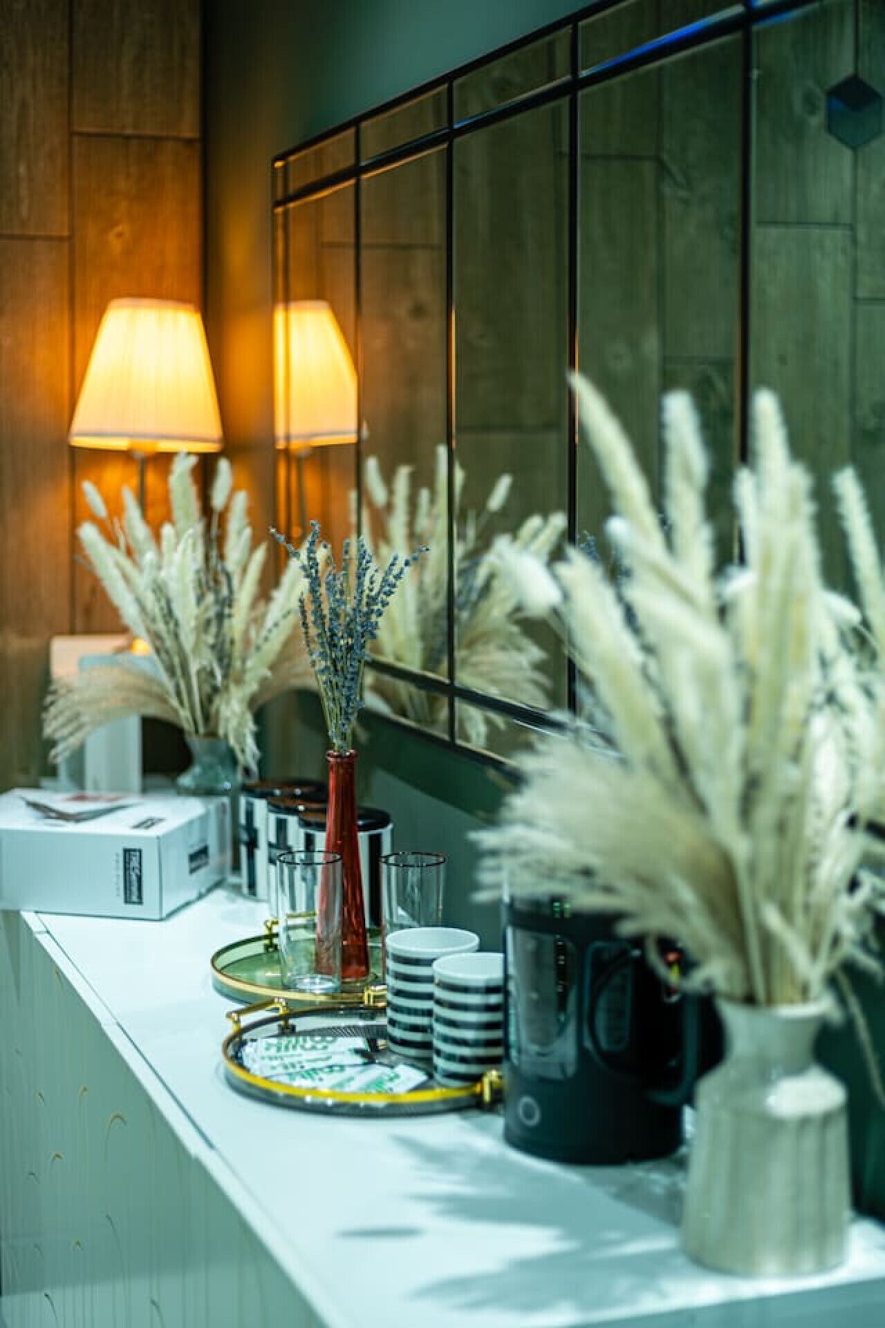 A modern bathroom with a large mirror, a white countertop, and a vase with pampas grass.