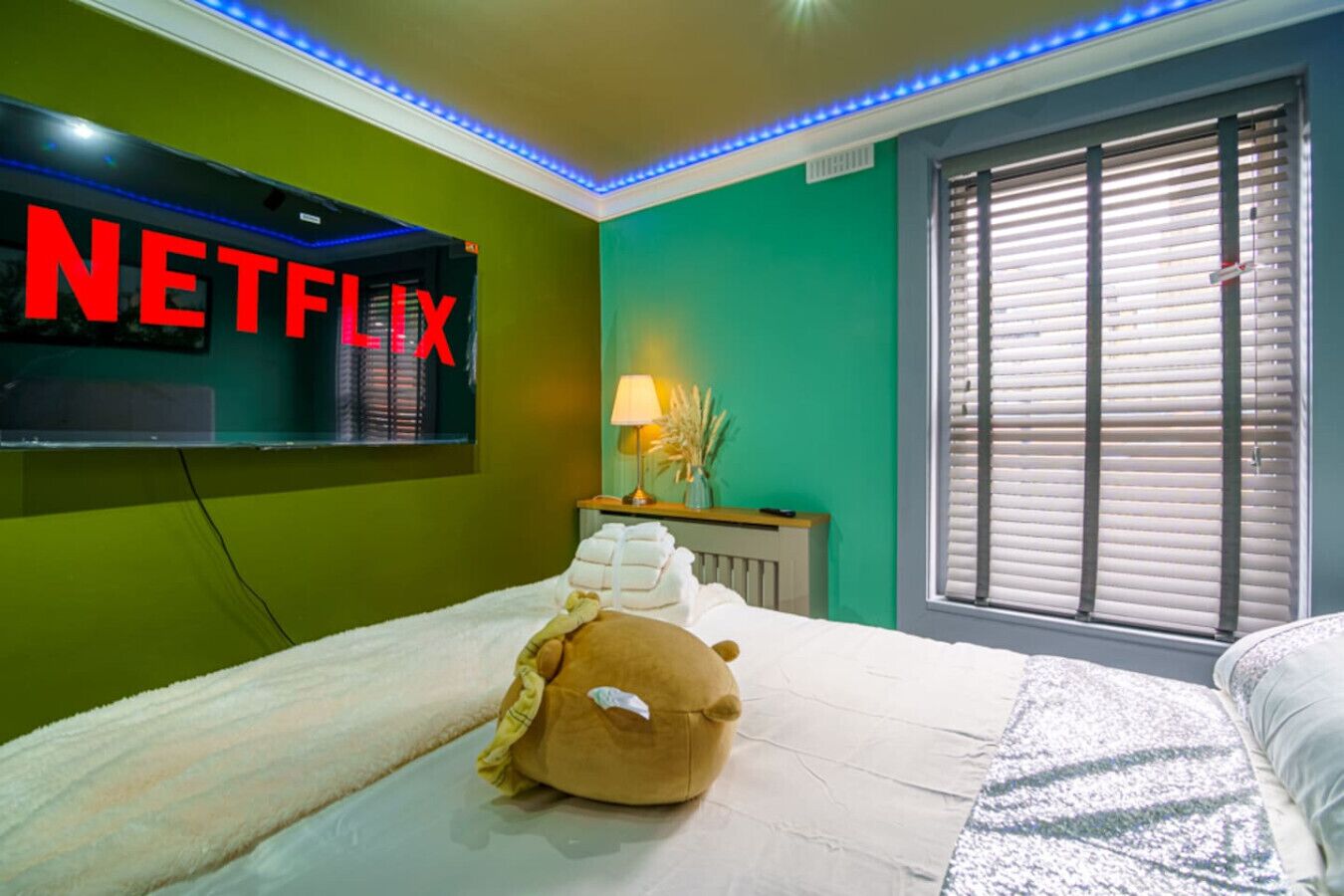 A hotel room with a bed, a wall-mounted TV displaying 'NETFLIX', a window with blinds, and a bedside table with a lamp and decorative items.