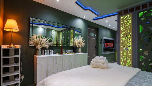 Une chambre d’hôtel moderne avec un grand lit, une coiffeuse avec un miroir, un logo Netflix et un élément de décoration séparant l’espace, illuminé de lumière verte.
