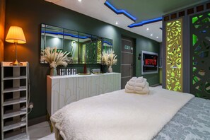 Une chambre d’hôtel moderne avec un grand lit, une coiffeuse avec un miroir, un logo Netflix et un élément de décoration séparant l’espace, illuminé de lumière verte.