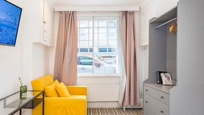 Quarto compacto com sofá amarelo, janela com cortinas, radiador e guarda-roupa cinzento, além de um quadro na parede.