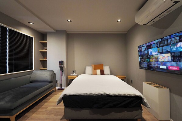 A modern hotel room with a bed, a sofa, a TV wall, and a shelf unit.