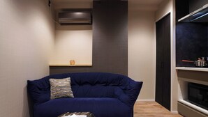 A compact living space with a dark blue sofa, a small table with plates, a wall-mounted air conditioner, and a microwave on a shelf.