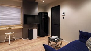 A compact living space with a flat-screen TV, a black refrigerator, a small table with a remote control, a white stool, and a blue sofa.