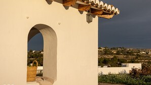A building with a white exterior, a wooden balcony, and a view of a landscape with houses and vegetation.