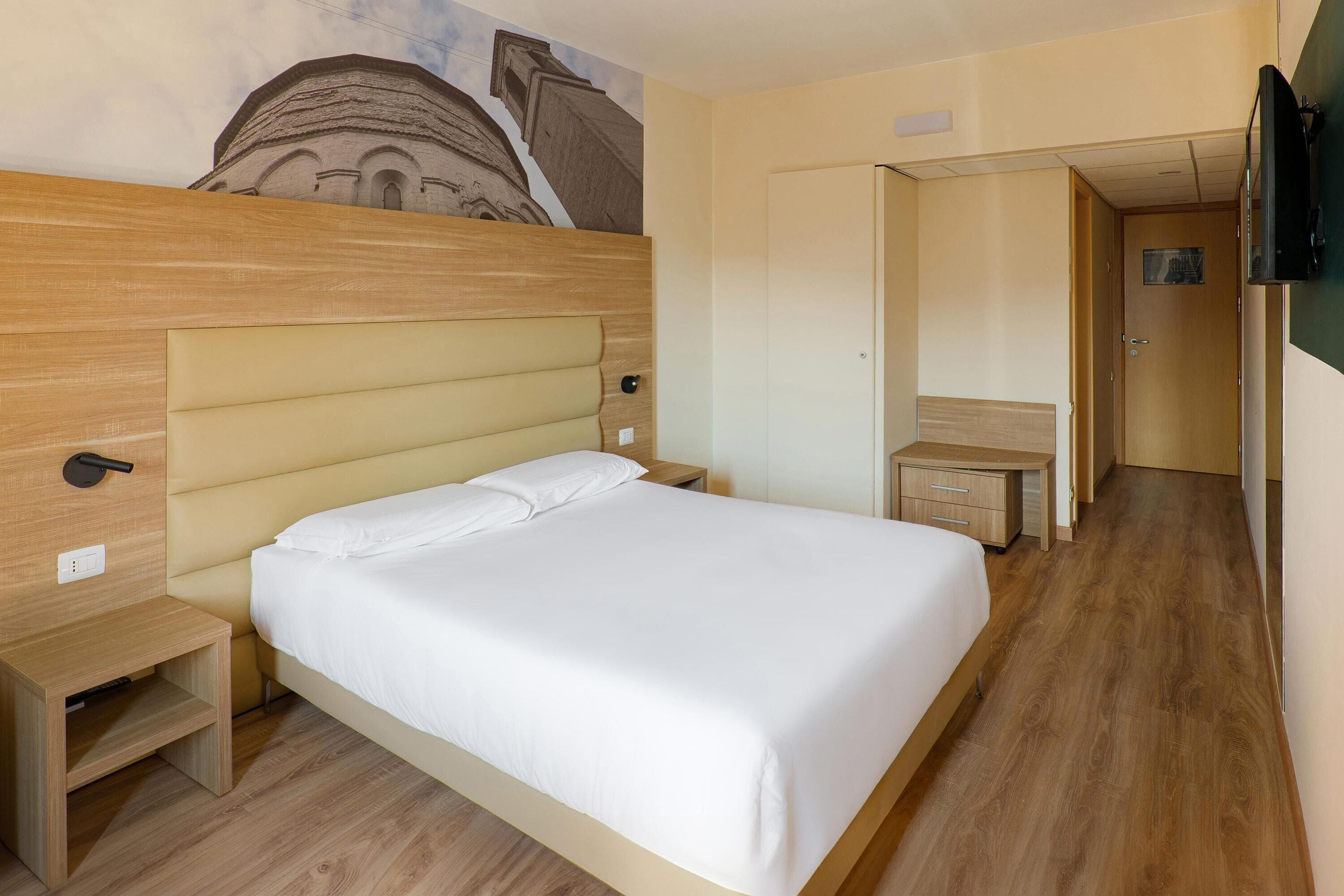 Une chambre d’hôtel avec un grand lit, une tête de lit en bois, des tables de chevet, un petit bureau et un téléviseur fixé au mur.