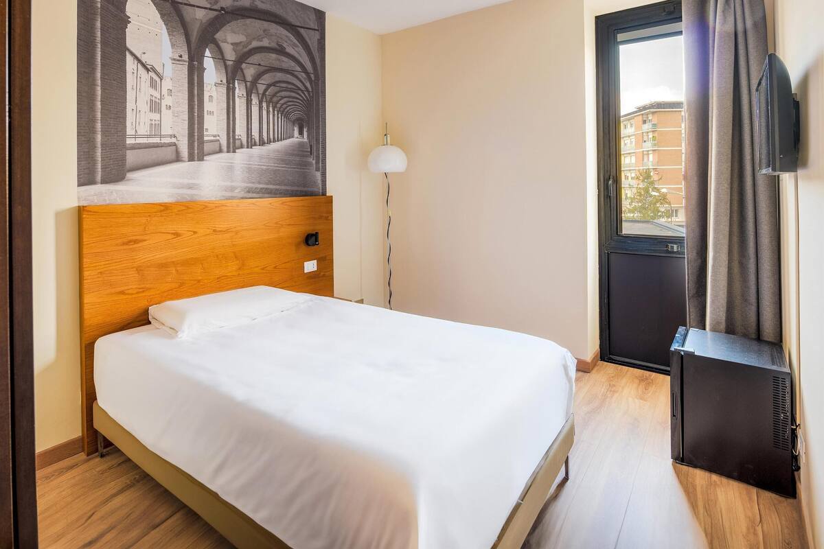 Ein Hotelzimmer mit Bett mit Holz-Kopfteil, einem Flachbildfernseher, einem Bild von einem architektonischen Korridor und Blick auf Gebäude durch das Fenster.