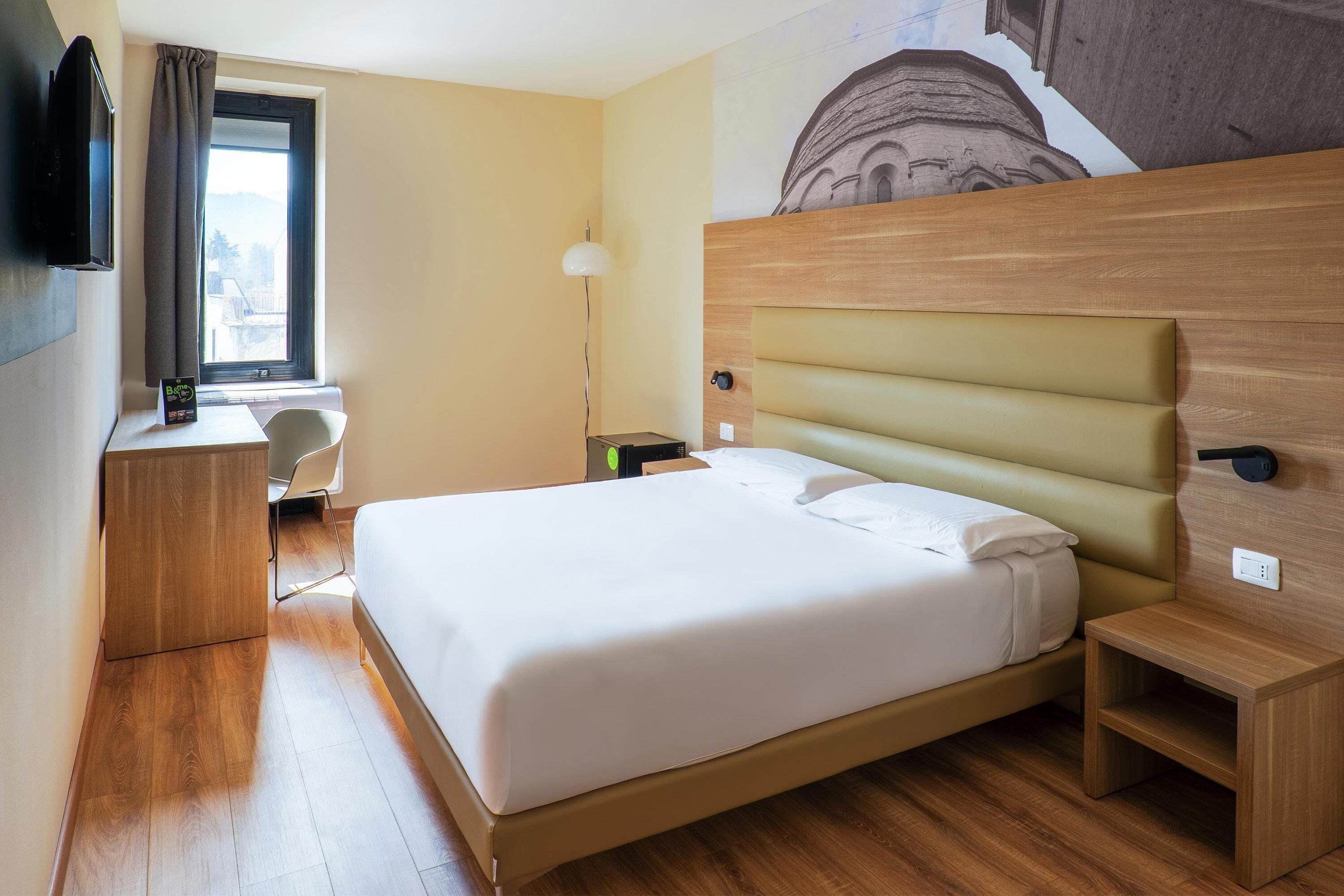 Una habitación de hotel con una cama grande, un escritorio, una silla, un televisor y una ventana con cortinas.
