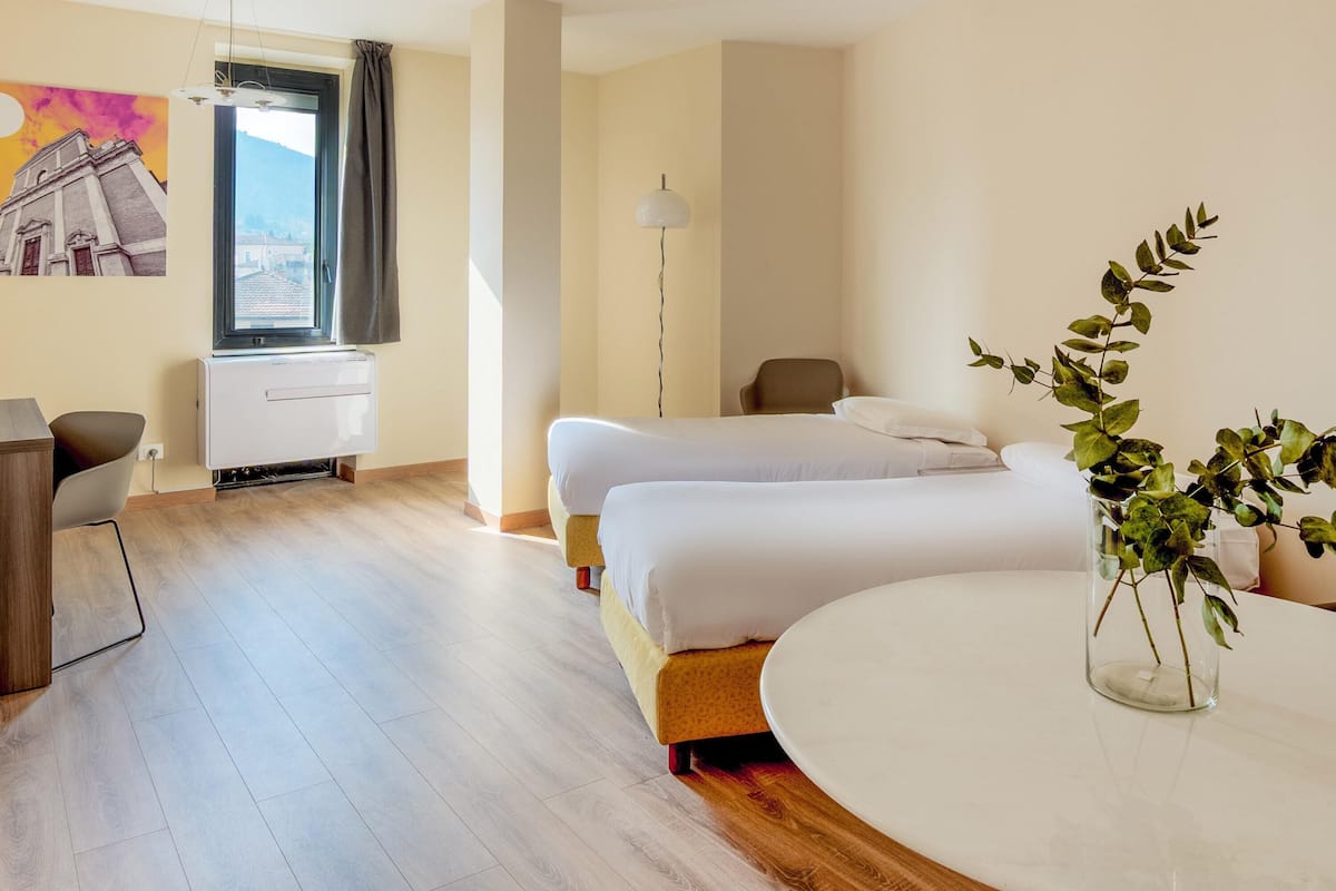 Ein Hotelzimmer mit zwei Betten, einem Schreibtisch, einem Stuhl, einem runden Tisch, einer Lampe und einem Fenster mit Aussicht.