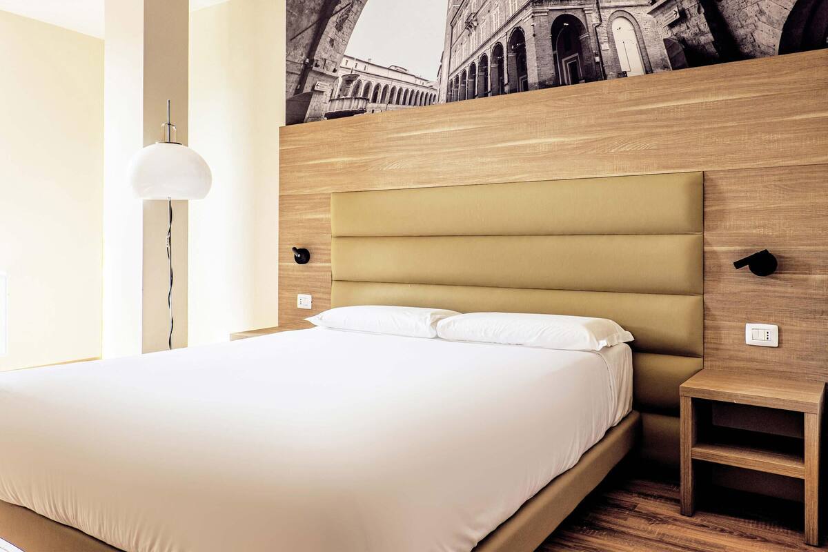 Ein modernes Hotelzimmer mit einem großen Bett, einem Kopfteil in heller Holzoptik, einer Wandlampe und einem kleinen Nachttisch.