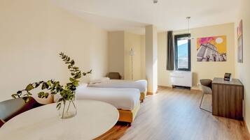 Habitación de hotel con dos camas, un escritorio, una silla, una mesa, un florero con planta, un cuadro y una ventana con vistas.