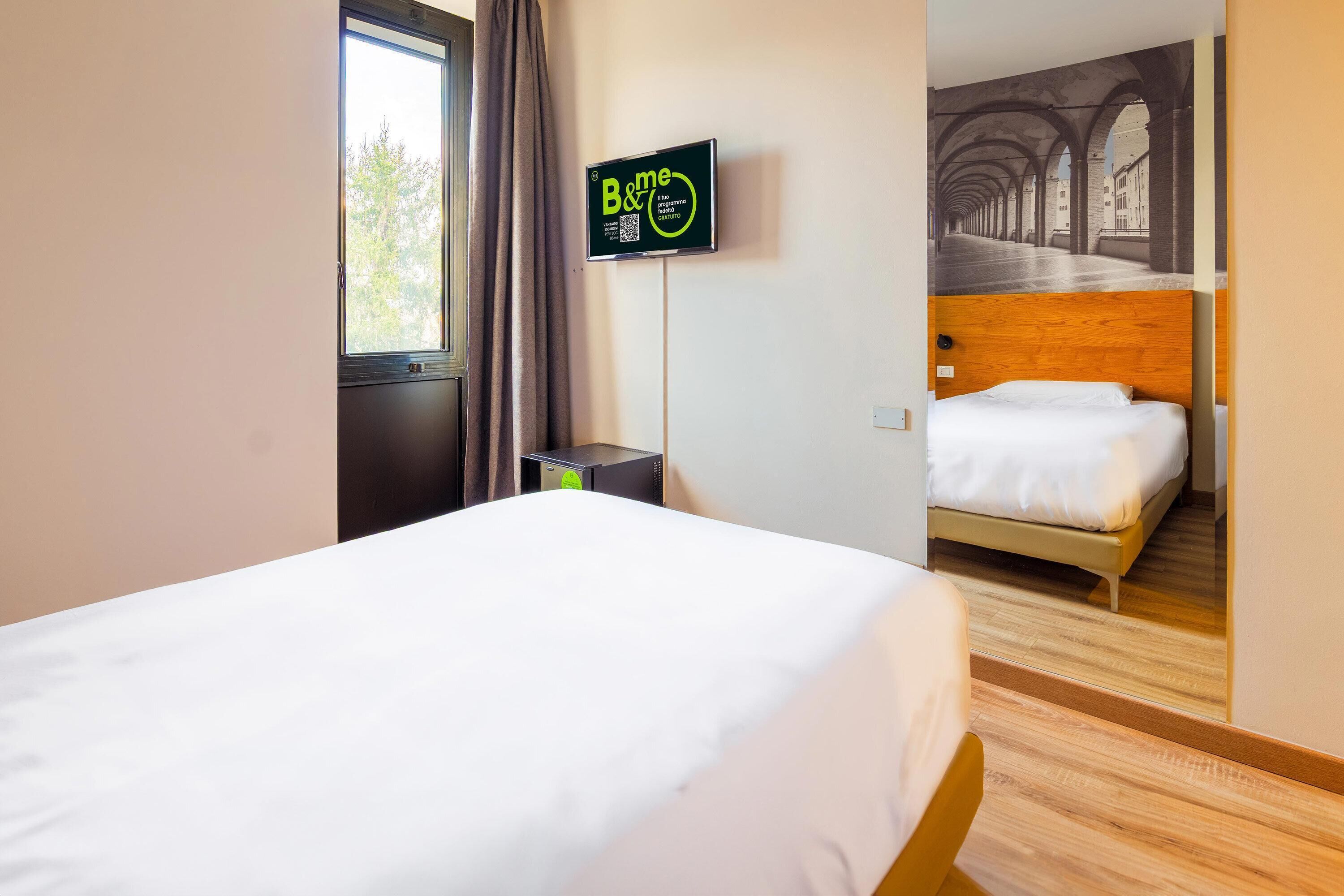 Une chambre d’hôtel avec un grand lit, un téléviseur fixé au mur et une vue sur un couloir extérieur par une fenêtre ouverte.