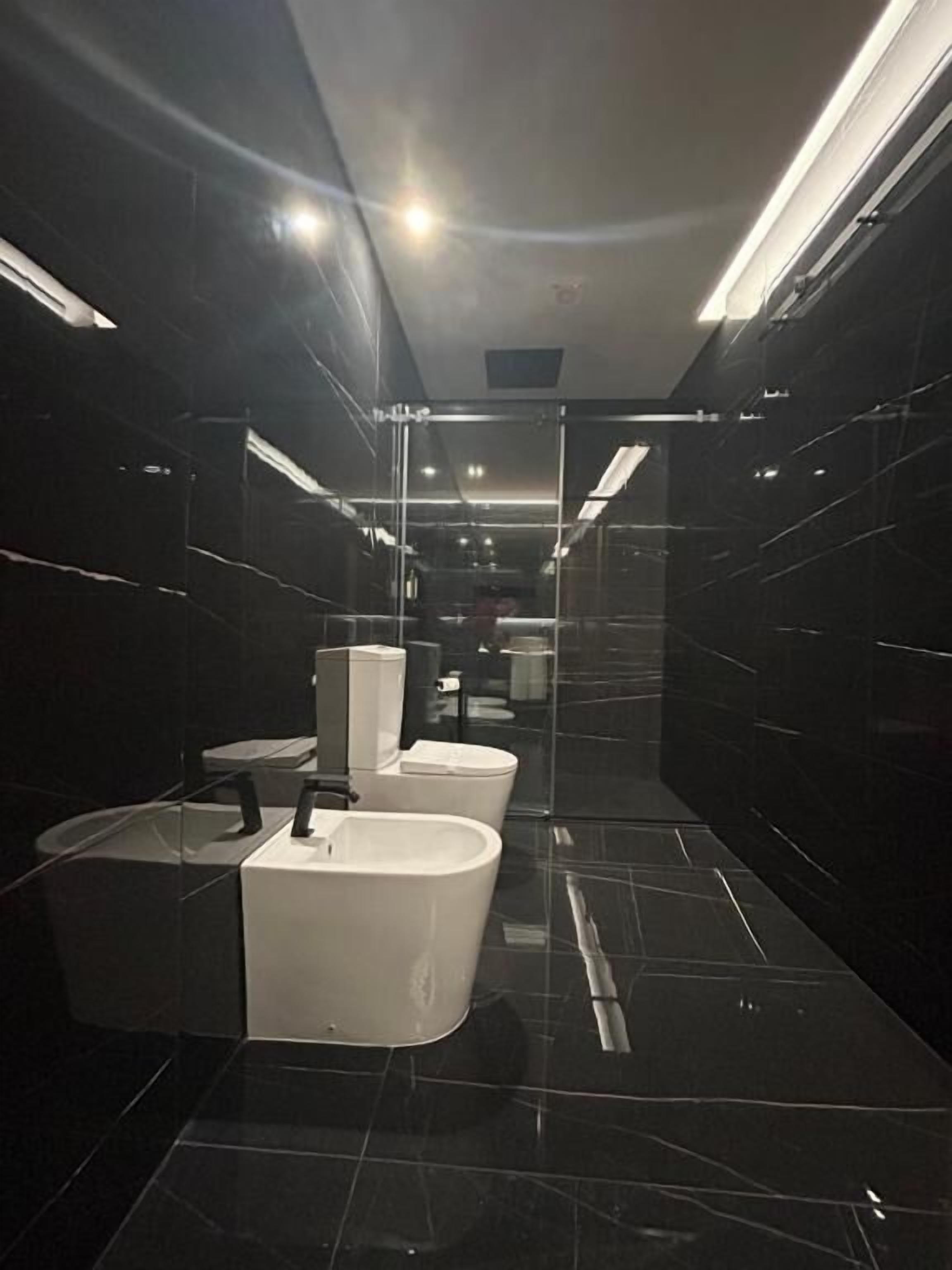 Un baño moderno con paredes de mármol negro, separador de vidrio y un lavamanos blanco central.