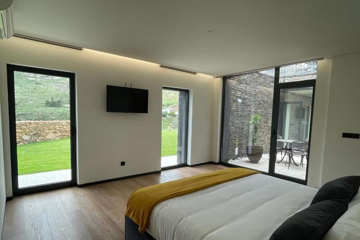 Ein modernes Schlafzimmer mit einem großen Bett, einem an der Wand montierten Fernseher und großen Fenstern mit Blick ins Freie.