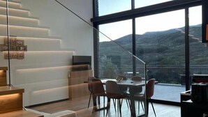 A modern interior with a staircase, dining area, and large windows offering a view of a mountainous landscape. - Empreendimento Serra (Covilhã)