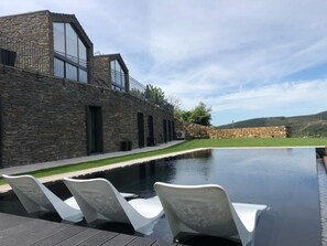 Un edificio moderno con fachada de piedra, una terraza en la azotea y una piscina con sillones de descanso.